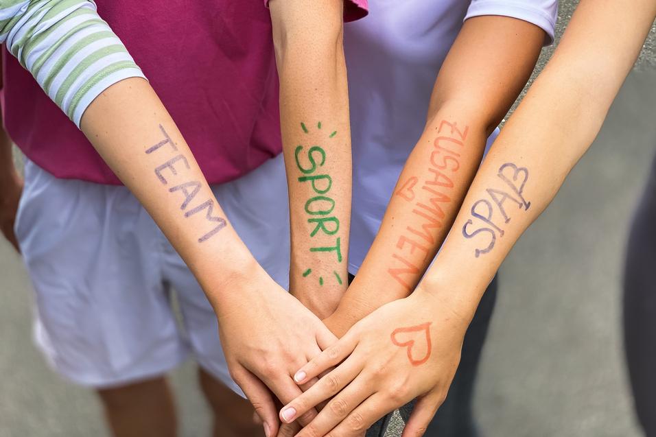 Vier Arme liegen übereinander, mit farbiger Schrift: „TEAM“, „SPORT“, „ZUSAMMEN“, „SPASS“ auf der Haut.