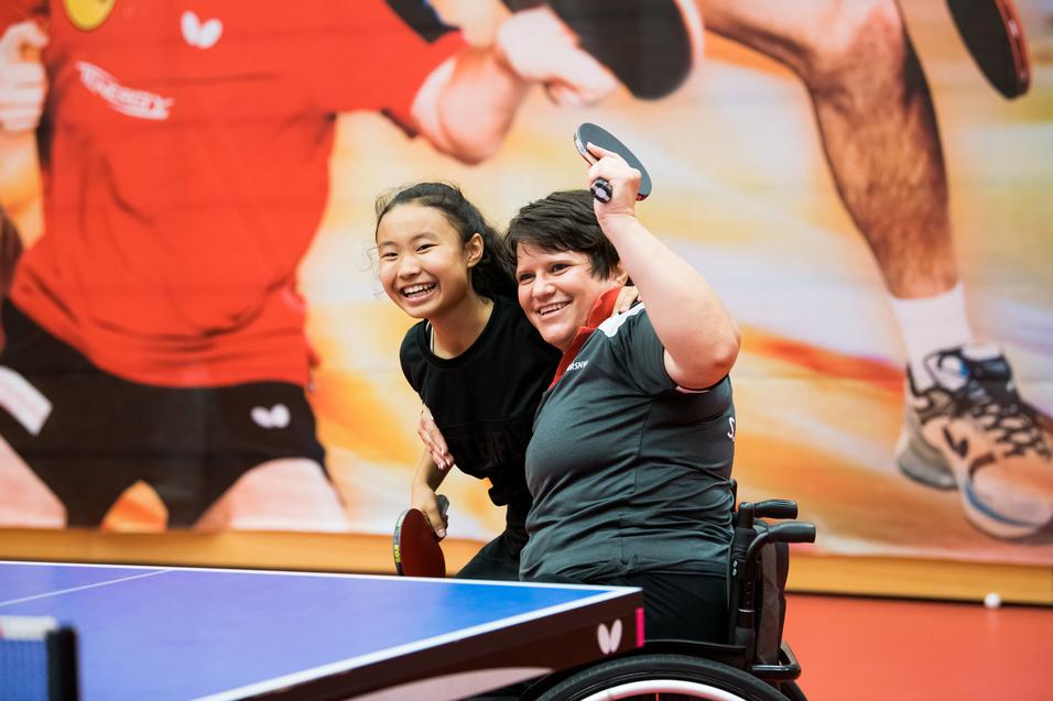 Zwei Frauen, eine im Rollstuhl, posieren fröhlich hinter einem Tischtennistisch mit Schlägern in der Hand.