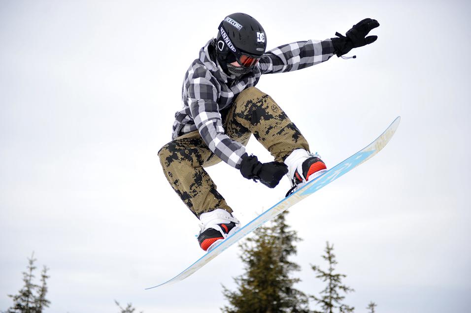 Snowboarder springt in der Luft, trägt eine karierte Jacke und camouflierte Hose, Hintergrund mit Nadelbäumen.